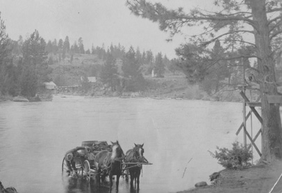 two horses stand in a river hitched to a wagon carrying water barrels that a man is filling from the river